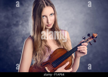 Attraktive junge Musikerin Stand halten ihre Geige im Arm gewiegt, beim Betrachten der Kameras mit einem Lächeln über b Stockfoto