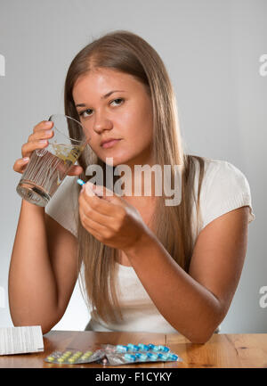 Hübsche junge Frau sitzt auf dem Tisch auf dem weißen Hintergrund, eine Pille und hält ein Glas Wasser zu Hause Stockfoto