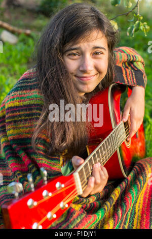 Teenager-Mädchen in Poncho spielt Gitarre beim Sitzen im Freien. Stockfoto