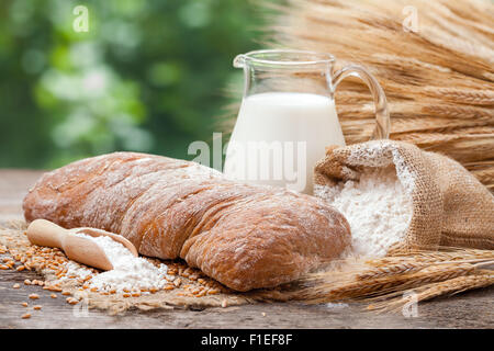 Frisches Brot, Krug mit Milch, Säckchen mit Mehl und Garbe Weizen auf Holztisch. Stockfoto