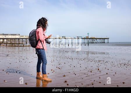 Einsame kaukasischen Brünette Reisende Frau mittleren Alters mit ihrem Handy am Strand in der Nähe von einem pier Stockfoto