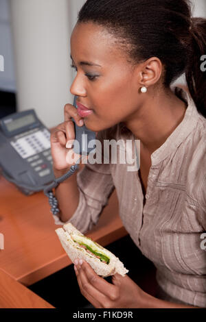 Weibliche Angestellte an der Rezeption ein Sandwich an ihrem Schreibtisch zu essen, während auf dem Telefon sprechen Stockfoto