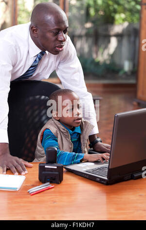 Ein afrikanischer Junge sitzt an seines Vaters Schreibtisch, auf ein Leptop zu spielen, während sein Vater über seine Schulter Uhren Stockfoto