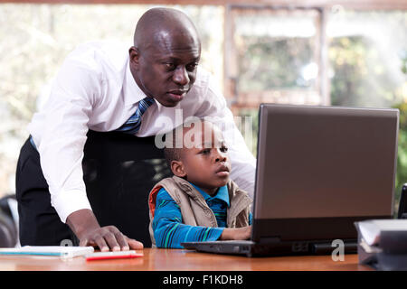 Ein afrikanischer Junge sitzt an seines Vaters Schreibtisch, auf ein Leptop zu spielen, während sein Vater über seine Schulter Uhren Stockfoto