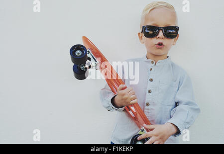 Entzückende Hipster kleiner Junge mit einem Skateboard tragen Sonnenbrillen entlehnt seine Mama oder Papa sich gegen eine Holzwand Giv Stockfoto