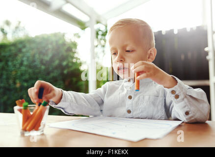 Süße kleine blonde Junge Auswahl eines farbigen Wax crayon aus einer Sammlung in einem Glasbehälter, wie er sich vorbereitet zu beginnen Zeichnung o Stockfoto