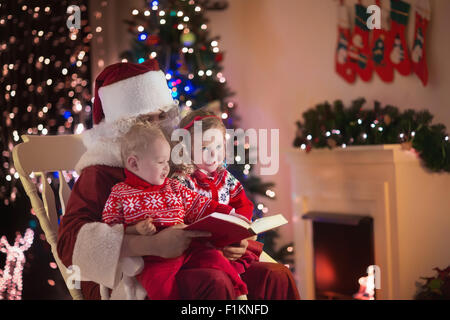 Santa Claus und Kinder öffnen präsentiert am Kamin. Kinder und Vater in Santa Kostüm und Bart öffnen Weihnachtsgeschenke Stockfoto