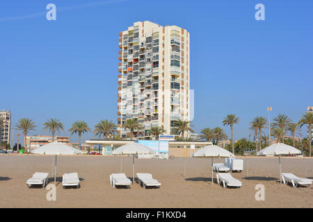 Liegestühle und Sonnenschirme am Strand von Playa San Juan mit einem Hochhaus Wohnblock hinter, Alicante, Spanien. Stockfoto