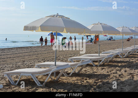 Sonnenliegen und Sonnenschirme aufgereiht am Strand in San Juan Playa, Alicante, mit Menschen in den frühen Morgenstunden am Strand ankommen. Stockfoto