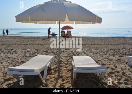 Liegestühle und Sonnenschirm am Strand von San Juan Playa, Alicante, mit Menschen zu Fuß an der Küste in den frühen Morgenstunden. Stockfoto