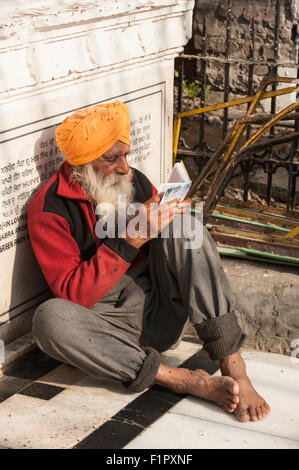 Amritsar, Punjab, Indien.  Der Goldene Tempel - Harmandir Sahib - mit einem alten grau bärtiger Sikh Mann in einem orangefarbenen Turban sitzen auf dem Boden lesen aus einem heiligen Buch, barfuß. Stockfoto