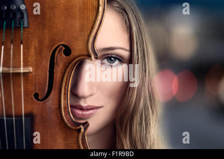 Dicht, ziemlich junge Frau spähen hinter Violine Instrument vor Lichter am Abend oder Nacht Stockfoto