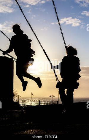 Silhouette von zwei weiblichen Kinder spielen auf Schaukeln vor blauem Himmel mit Sonne beginnen, legen Sie Stockfoto