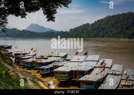 Langbooten angedockt am Bootsanleger auf dem Mekong in Luang Prabang Laos. Berge in Ferne in dieser Baum-gerahmte Ansicht sichtbar Stockfoto