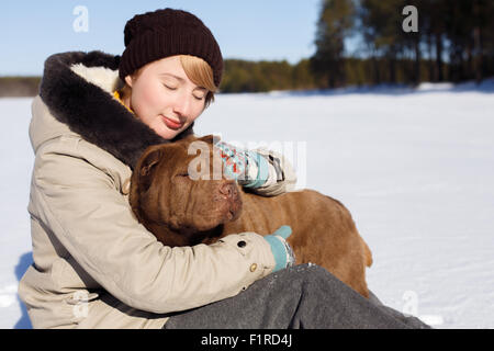 Frau umarmt ihren Shar pei sitzend auf dem verschneiten Feld in der Nähe von einem Fichtenwald an einem sonnigen Tag. Winter Unterkunftstyp Konzept. Hundetraining. Stockfoto