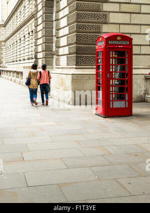 Einzelne rote Telefonzelle in London in leuchtend rote Farbe und mit entsättigten Farben mit zwei unbekannten Touristen vorbeigehen Stockfoto