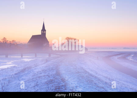 Eine Kirche in eine gefrorene Winterlandschaft in den Niederlanden. Bei Sonnenaufgang an einem schönen nebligen Morgen fotografiert. Stockfoto