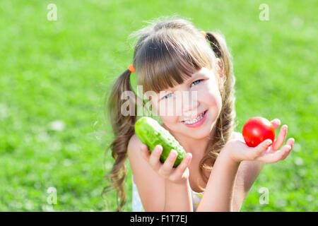 Glückliches kleines Mädchen sitzen auf dem Rasen mit Gemüse am Tageszeit. Konzept von gesunden Lebensmitteln. Stockfoto