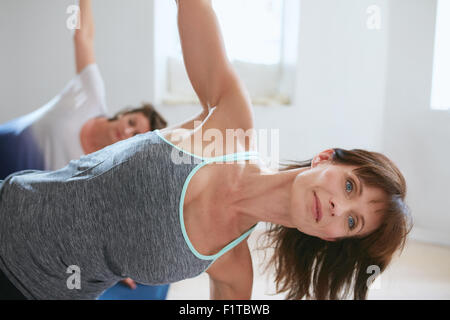 Porträt von Reife Frau tun erstreckt sich in einem Fitnessstudio. Fitness-Trainer mit Studenten im Hintergrund Ardha Chandrasana zu tun. Halbmond Stockfoto