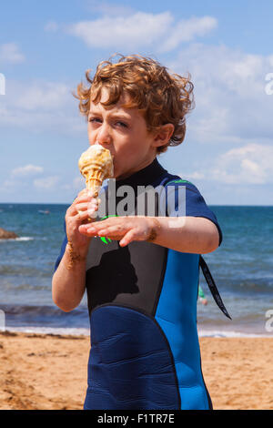 Sechs Jahre alter Junge, Essen eine Eis trägt einen Neoprenanzug auf Hope Cove, Devon, England, Vereinigtes Königreich. Stockfoto