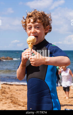 Sechs Jahre alter Junge, Essen eine Eis trägt einen Neoprenanzug auf Hope Cove, Devon, England, Vereinigtes Königreich. Stockfoto