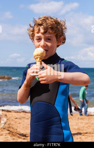 Sechs Jahre alter Junge, Essen eine Eis trägt einen Neoprenanzug auf Hope Cove, Devon, England, Vereinigtes Königreich. Stockfoto