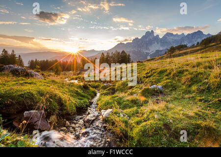 Die Sonne geht hinter den Bergen und wirft das letzte Licht auf eine hohe Weide mit einem kleinen Gebirgsbach. Stockfoto