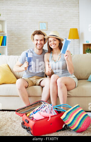 Glückliches Paar zeigt Flugtickets sitzend auf Sofa mit Koffer, die Vorbereitungen für den Flug in der Nähe von Stockfoto