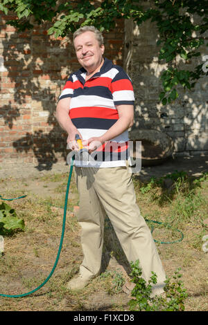 In voller Länge Portrait lächelnd reifen Mann, der den Garten des Grundstückes mit Gartenschlauch mit Spritzpistole drauf Gießen ist. Stockfoto