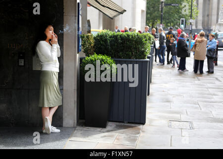 London-2015: Ein Büroangestellter findet eine Zigarettenpause außerhalb ihrer Bürogebäude welche Fronten auf einer belebten Straße. Sie verwenden ihr Handy Stockfoto