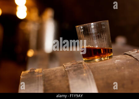 Glas gefüllt mit Whisky, ordentlich ohne Eis, stehend auf einem Holzfass in Brennerei mit Fässern im Hintergrund. Stockfoto