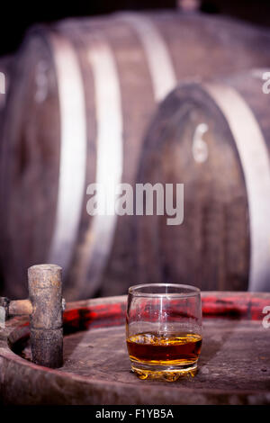 Glas gefüllt mit Whisky, ordentlich ohne Eis, stehend auf einem Holzfass in Brennerei mit Fässern im Hintergrund. Stockfoto