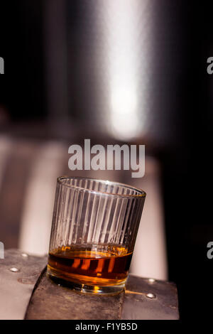 Glas gefüllt mit Whisky, ordentlich ohne Eis, stehend auf einem Holzfass in Brennerei mit Fässern im Hintergrund. Stockfoto