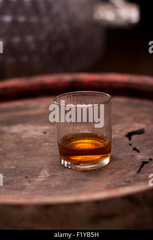 Glas gefüllt mit Whisky, ordentlich ohne Eis, stehend auf einem Holzfass in Brennerei mit Fässern im Hintergrund. Stockfoto