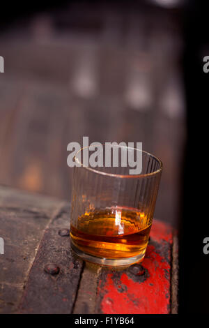 Glas gefüllt mit Whisky, ordentlich ohne Eis, stehend auf einem Holzfass in Brennerei mit Fässern im Hintergrund. Stockfoto