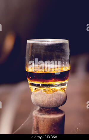 Glas gefüllt mit Whisky, ordentlich ohne Eis, stehend auf einem Holzfass in Brennerei mit Fässern im Hintergrund. Stockfoto