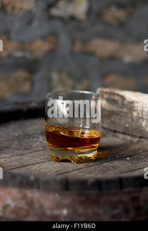 Glas gefüllt mit Whisky, ordentlich ohne Eis, stehend auf einem Holzfass in Brennerei mit Fässern im Hintergrund. Stockfoto