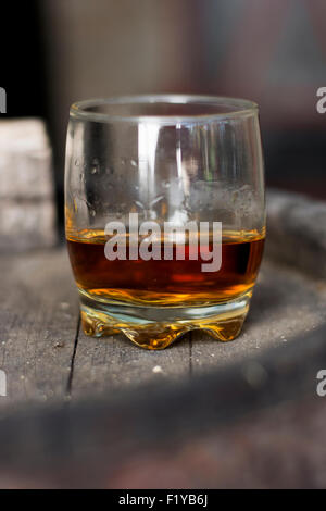 Glas gefüllt mit Whisky, ordentlich ohne Eis, stehend auf einem Holzfass in Brennerei mit Fässern im Hintergrund. Stockfoto