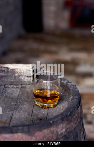 Glas gefüllt mit Whisky, ordentlich ohne Eis, stehend auf einem Holzfass in Brennerei mit Fässern im Hintergrund. Stockfoto