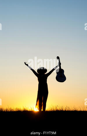 Teenager-Mädchen stand hält eine Gitarre mit ihren Armen in der Luft bei Sonnenuntergang. Silhouette Stockfoto