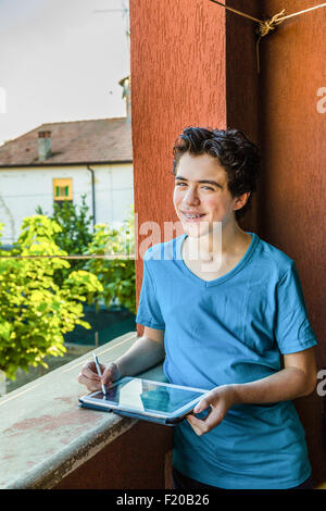 Latin Boy lächelt zeigt Klammern, während er einen Tablet-PC mit einer roten Grunge-Wand hält und grüne als Hintergrund Blätter Stockfoto