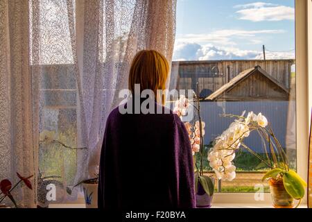 Frau mit Blick auf landwirtschaftliche Gebäude aus dem Wohnzimmerfenster Stockfoto