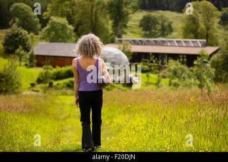 Rückansicht des Reife Frau, die Yoga-Matte zu Fuß in Eco-Lodge-Feld Stockfoto