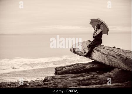 Frau mit Regenschirm auf große Treibholz Baumstamm am Strand sitzen Stockfoto