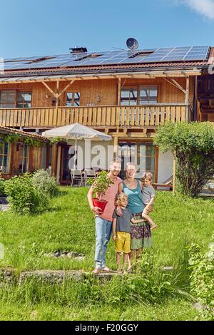 Porträt der jungen Familie, Topfpflanze halten, stand vor Haus mit holzverkleideten Solardach Stockfoto