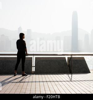 Silhouette Seitenansicht der junge Frau auf der ständigen Suche aus über Wasser im Skyline, Hong Kong, China Stockfoto