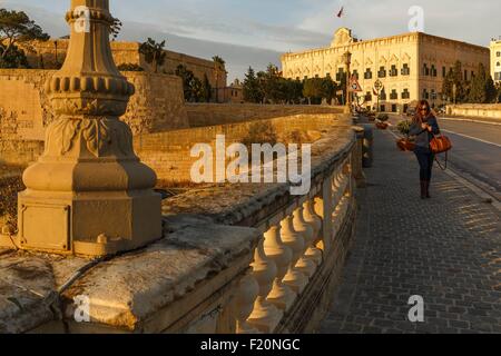 Malta, Valletta, Weltkulturerbe der UNESCO, Stadtbild von einer jungen Frau zu Fuß auf einem Steg über die Stadtmauern und Befestigungsanlagen der Altstadt bei Sonnenuntergang Stockfoto