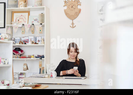 Eine ältere Frau sitzt mit einem Smartphone in einen Souvenirladen, einen kleinen Einzelhandel Geschäftsbetrieb. Stockfoto