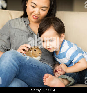 Lächelnde Frau sitzen auf einem Sofa mit ein Meerschweinchen sitzen auf dem Schoß, ihr kleiner Sohn beobachten. Stockfoto