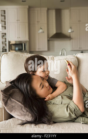 Frau liegt auf dem Sofa kuscheln mit ihrem kleinen Sohn und mit Blick auf ein Handy. Stockfoto